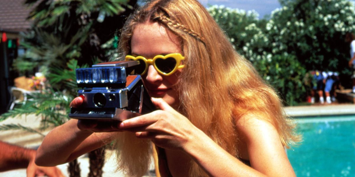 Blonde woman in yellow heart shaped sunglasses holding a camera, pool in the background with people standing around.